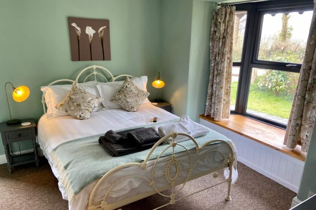 Une chambre à coucher comprenant un lit, deux tables de chevet, une fenêtre avec des rideaux et un tableau accroché au mur.