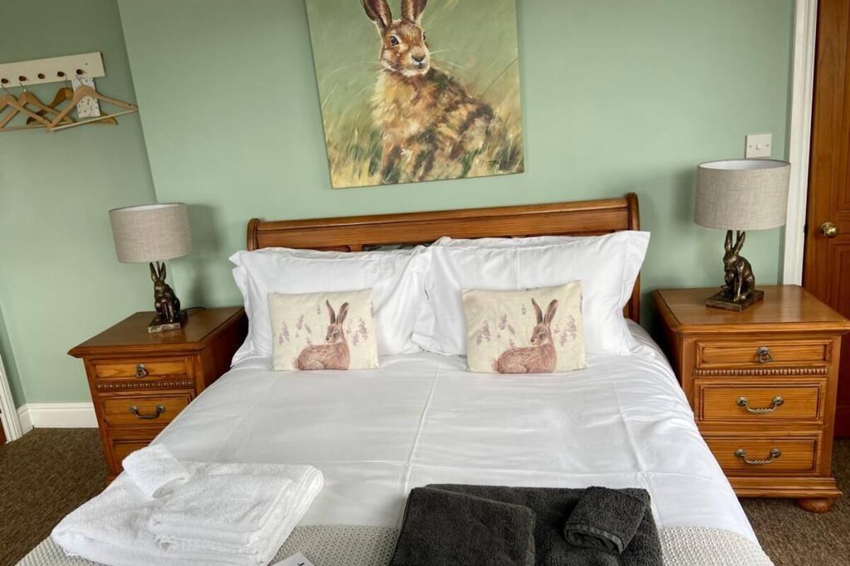 Un lit bien fait, avec des draps blancs et deux oreillers, une table de chevet en bois avec une lampe, et un tableau représentant un lapin accroché au mur.