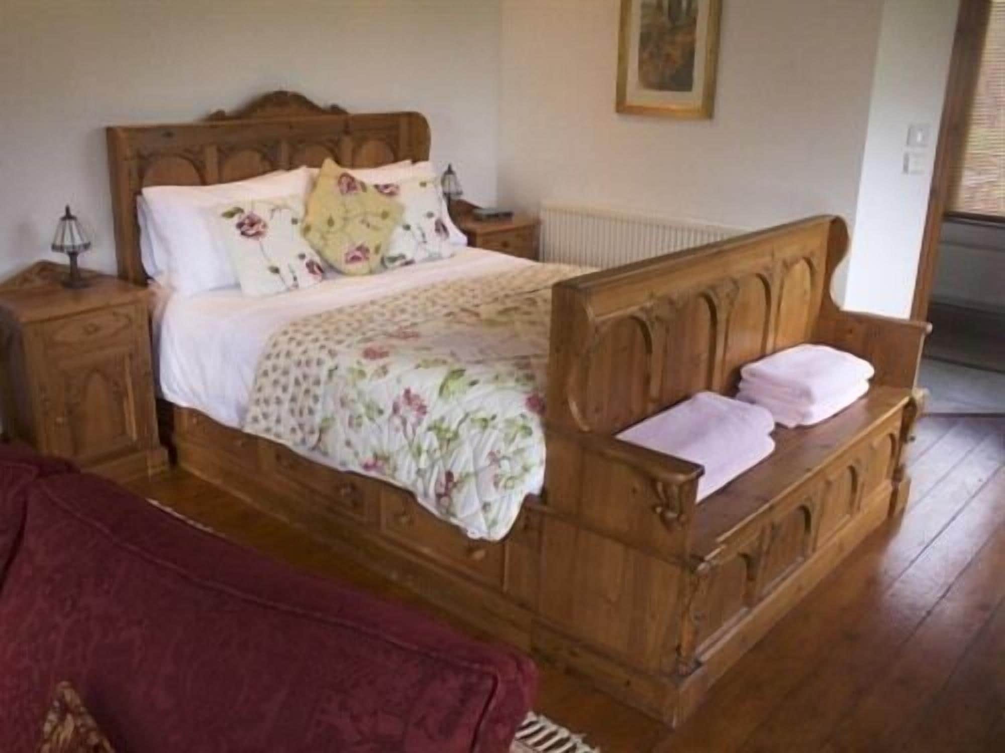 A wooden bed with a floral bedspread and pillows, a nightstand with a lamp, and a radiator on the wall.