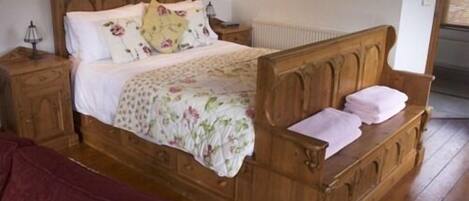 A wooden bed with a floral bedspread and pillows, a nightstand with a lamp, and a radiator on the wall.