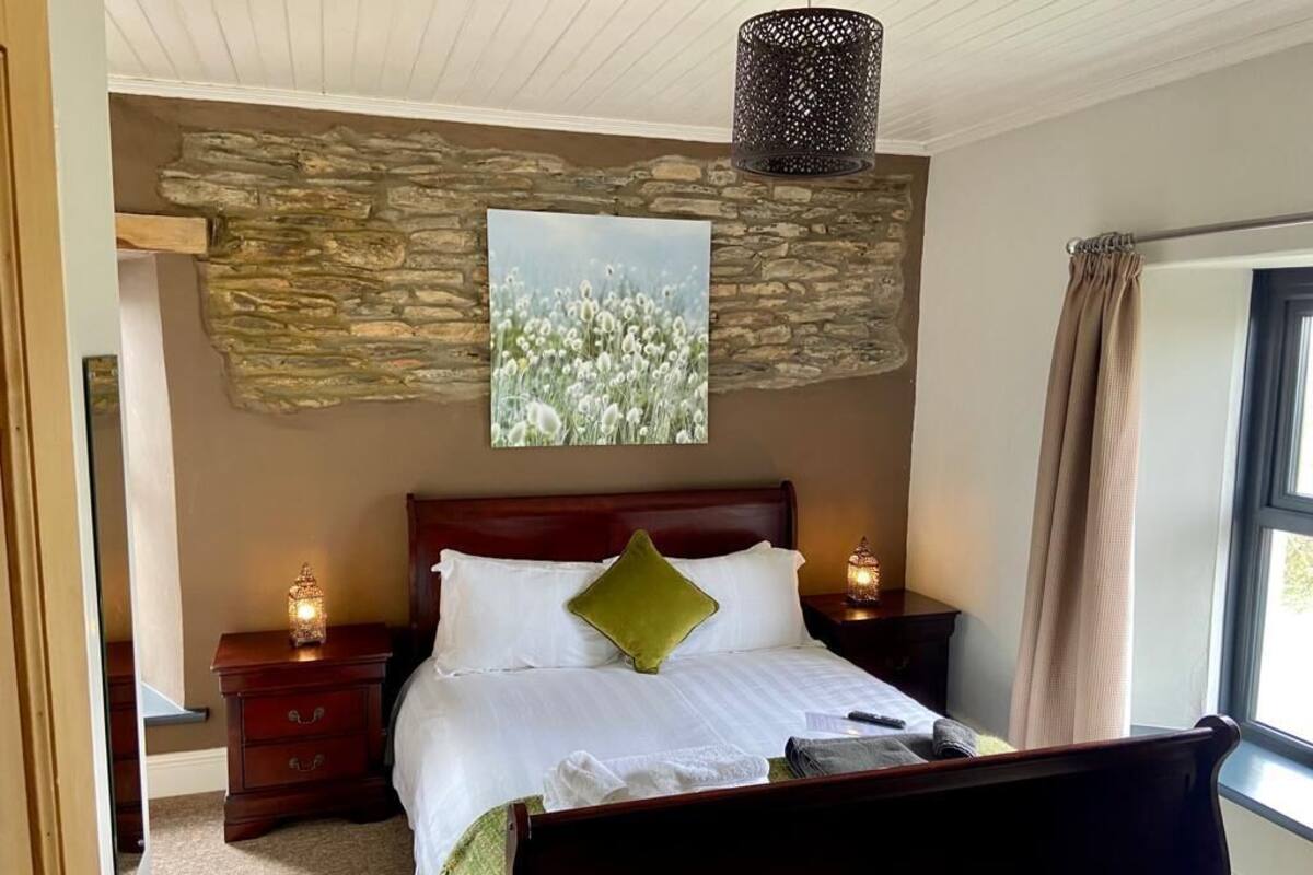 Une chambre avec un mur en pierre, un lit recouvert de draps blancs, un oreiller vert, deux tables de chevet avec des lampes et une fenêtre avec des rideaux.