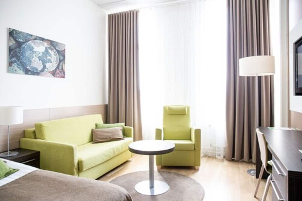 Une chambre d’hôtel moderne avec un canapé vert, une table ronde, un fauteuil vert et un téléviseur posé sur un meuble.