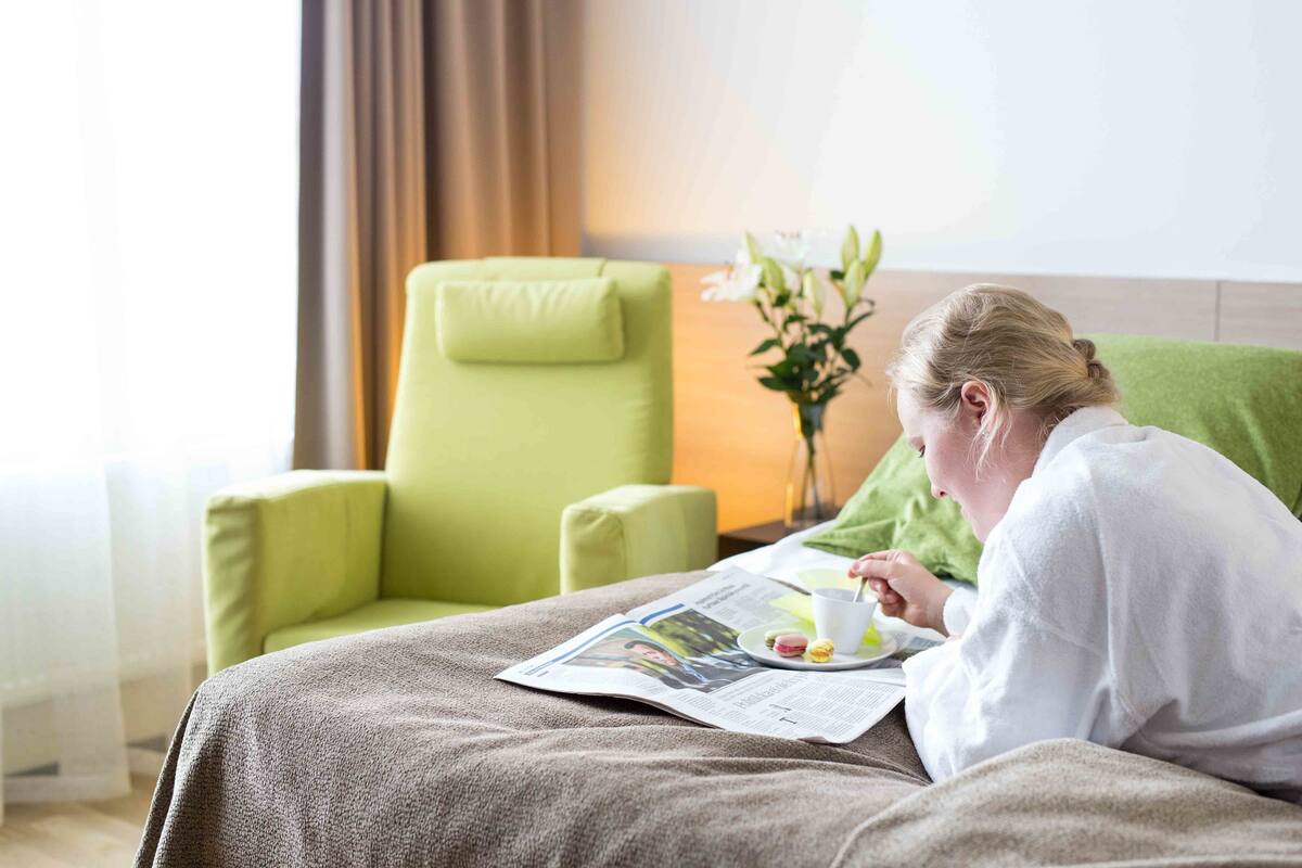 Une personne assise sur un lit, avec un journal, une tasse et une assiette contenant de la nourriture.