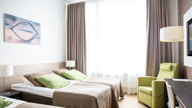 A hotel room with two beds, a green armchair, a desk, and a TV.