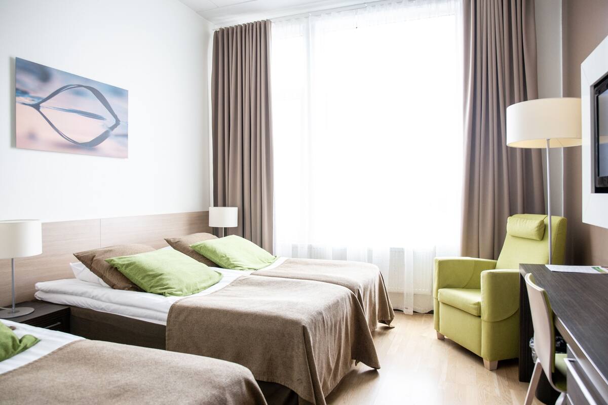 Une chambre d’hôtel avec deux lits, un fauteuil vert, un bureau et une télévision.