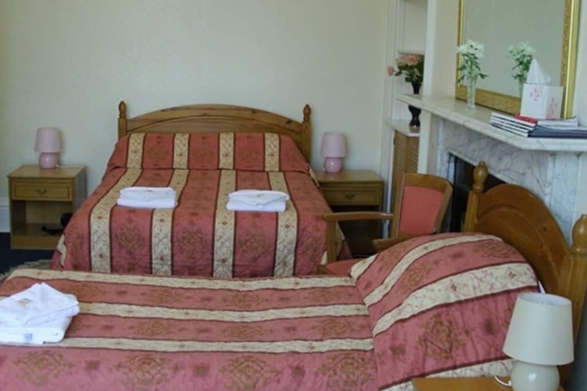 A neatly made bed with a patterned bedspread, two bedside tables with lamps, a chair, and a fireplace with a mirror above it.