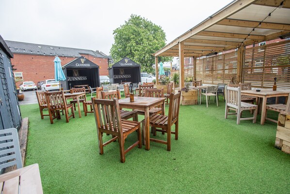 An outdoor seating area with wooden tables and chairs, two Guinness branded tents, and a brick building in the background.