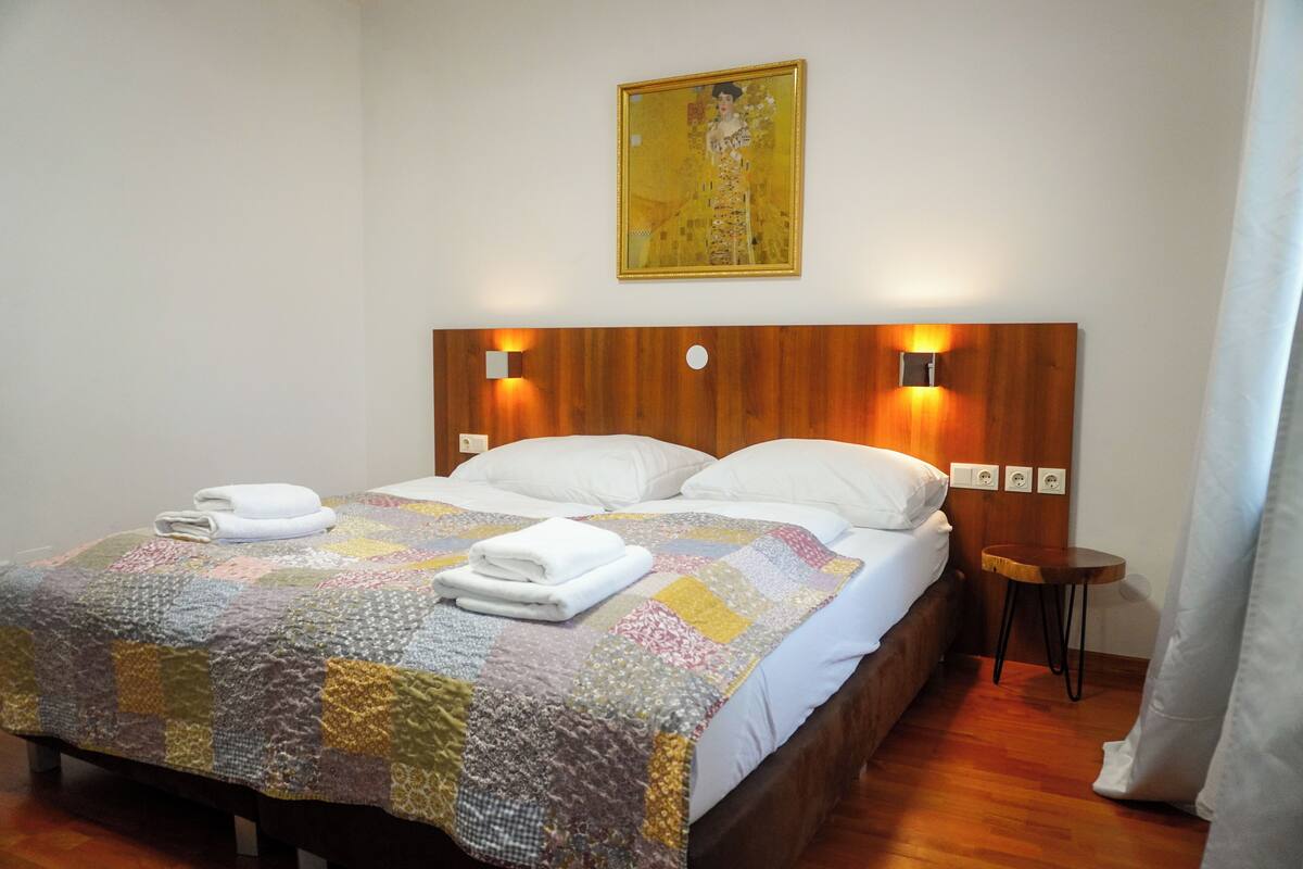 A neatly made bed with a colorful quilt, a wooden headboard with wall-mounted lights, and a painting above the bed.