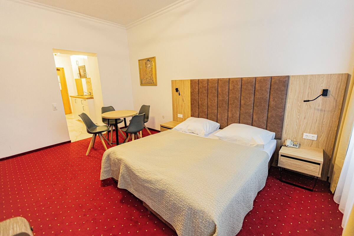 A hotel room with a large bed, a bedside table with a lamp, a chair, and a small table with a chair.