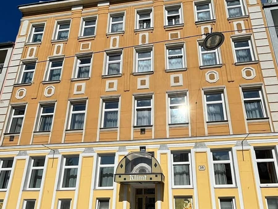 Ein mehrstöckiges Gebäude mit gelber Fassade, zahlreichen Fenstern und einem zentralen Eingang, der ein Schild mit der Aufschrift "Klimt" trägt.
