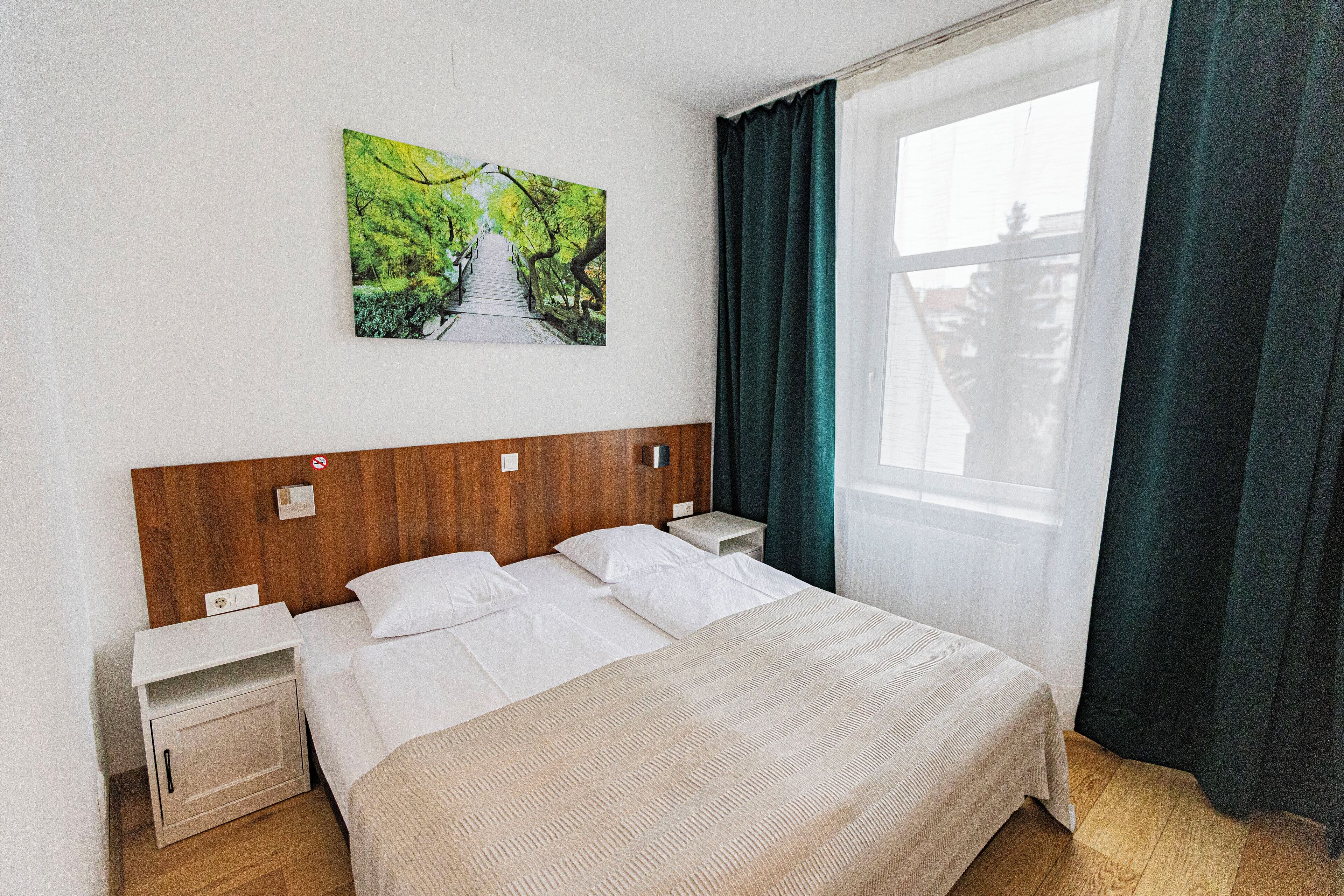Een hotelkamer met een houten hoofdeinde, een groot bed met witte lakens, een nachtkastje en een raam met gordijnen.