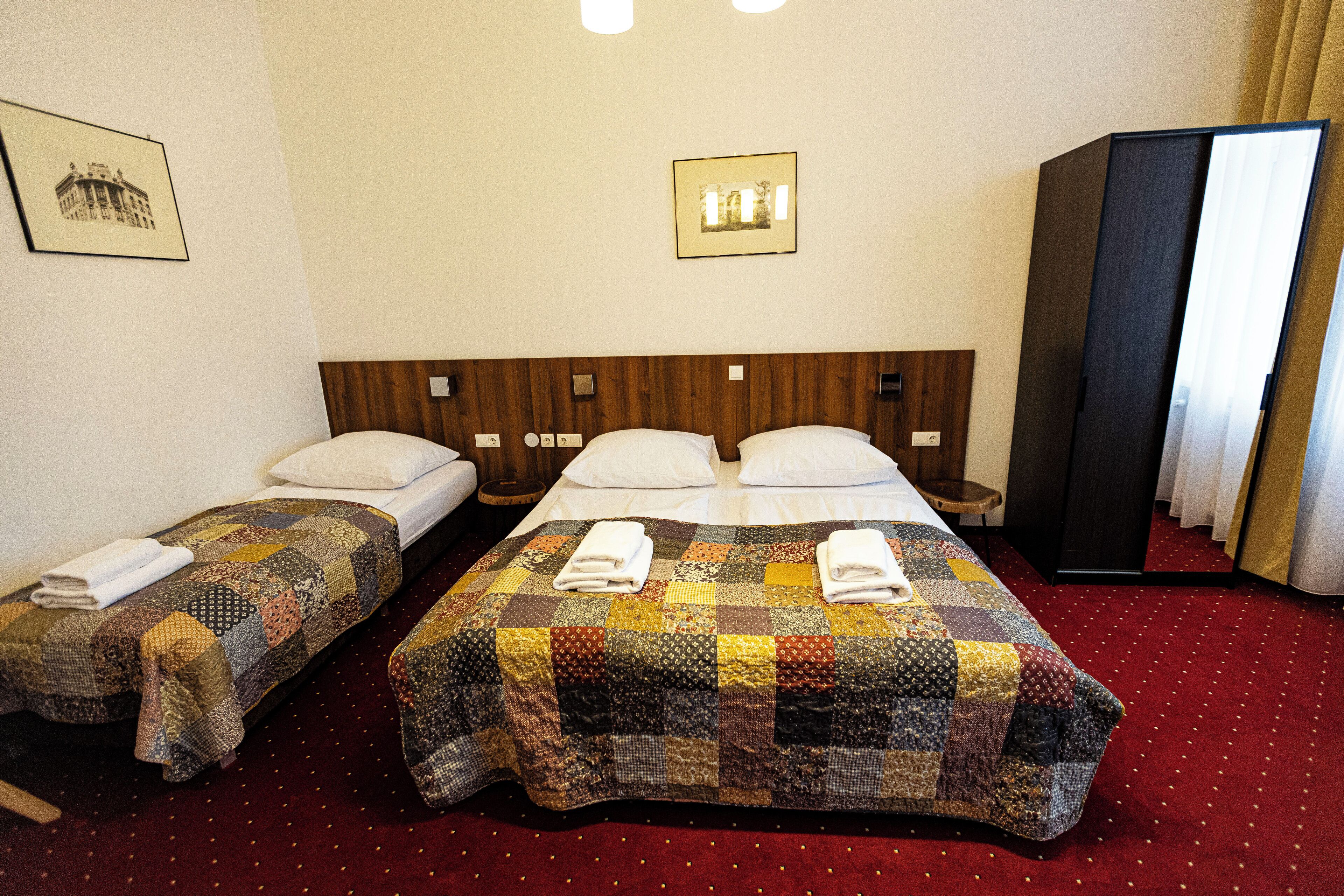 Une chambre d’hôtel avec deux lits, une tête de lit en bois, une grande armoire et une moquette rouge.