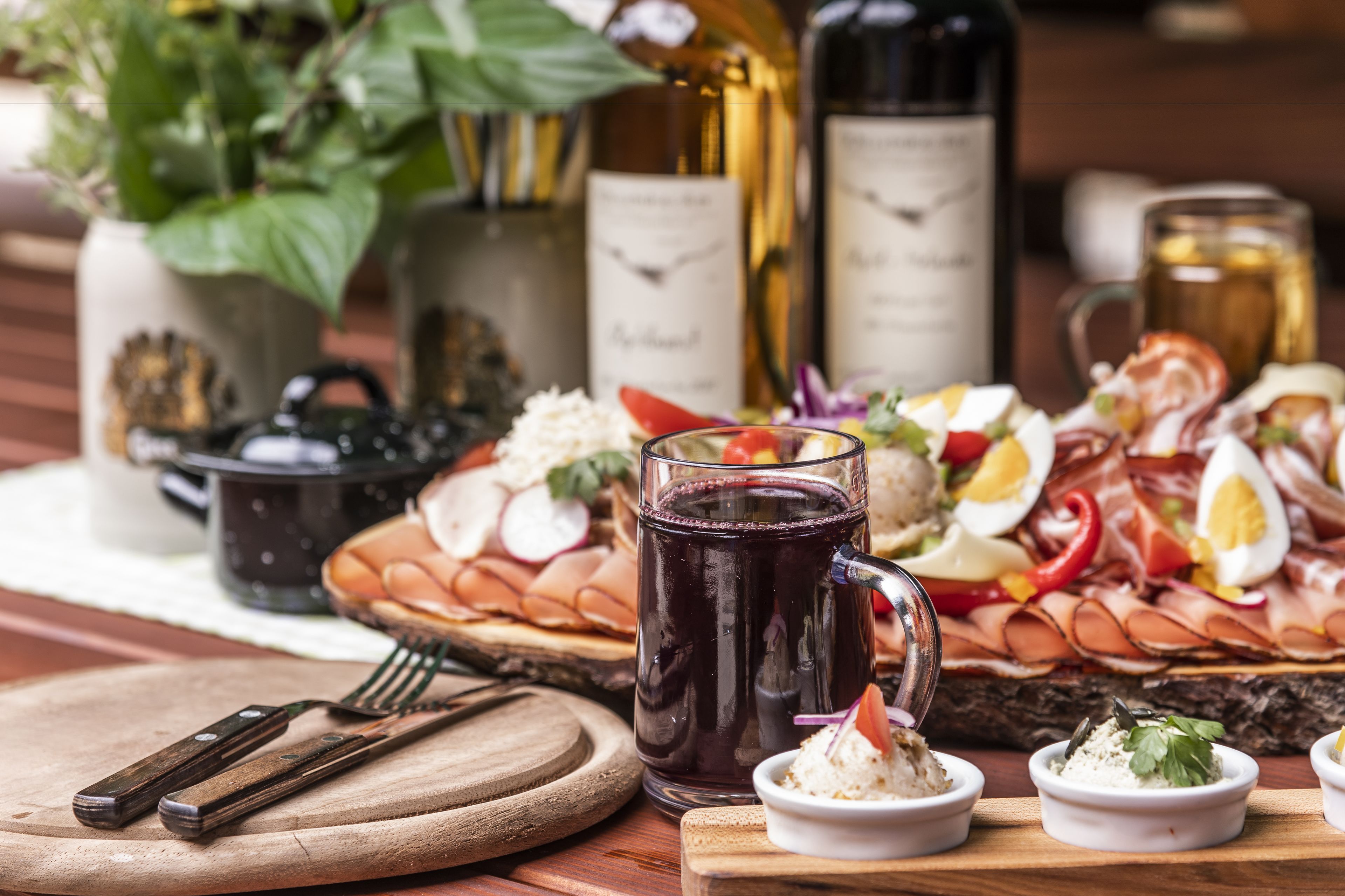 Una tabla de embutidos con diversos quesos, carnes y acompañamientos, junto con botellas de vino y una jarras de bebida.