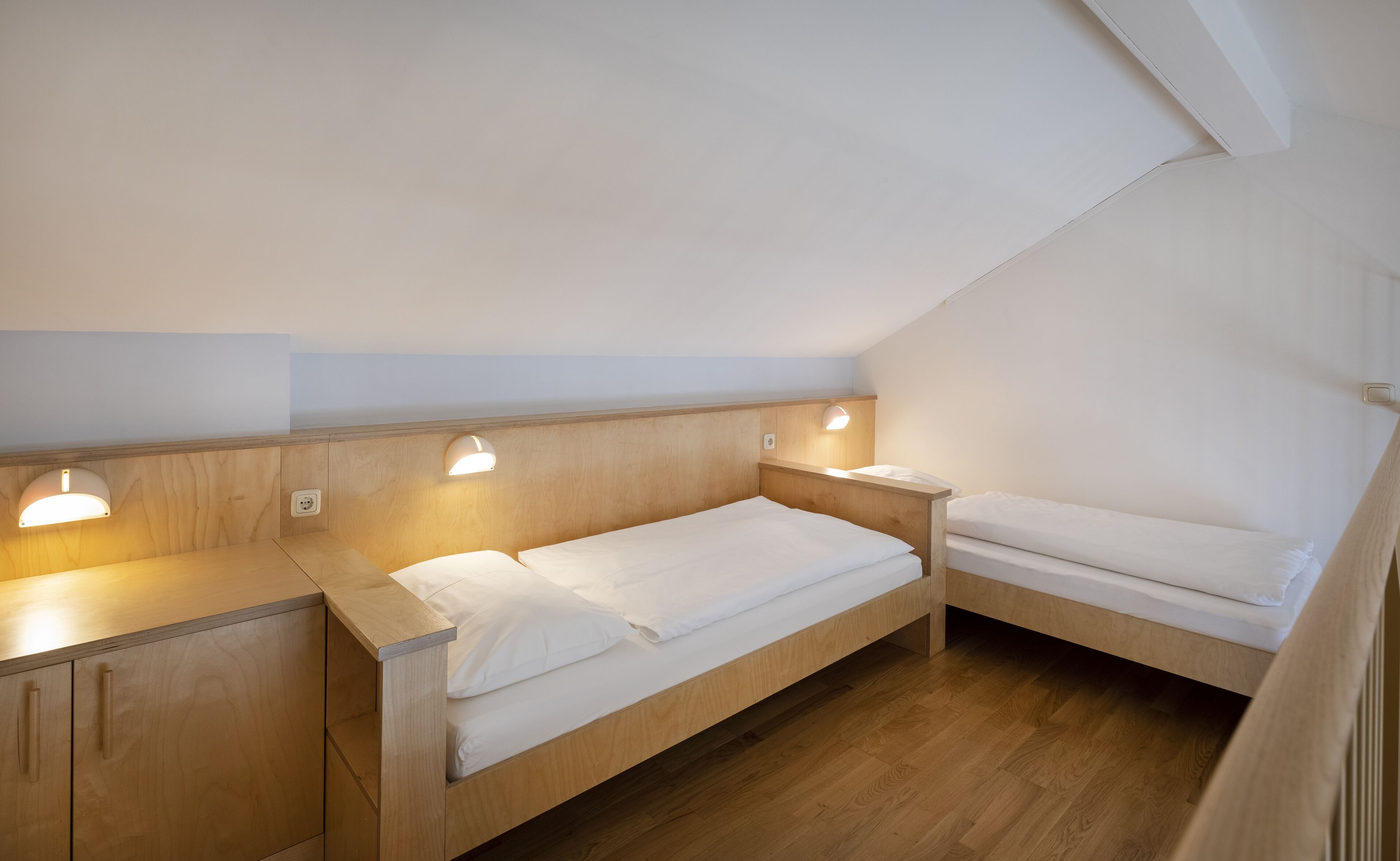 Una habitación pequeña y bien iluminada con dos literas de madera, cada una con una mesita de noche y una lámpara de lectura en la pared.