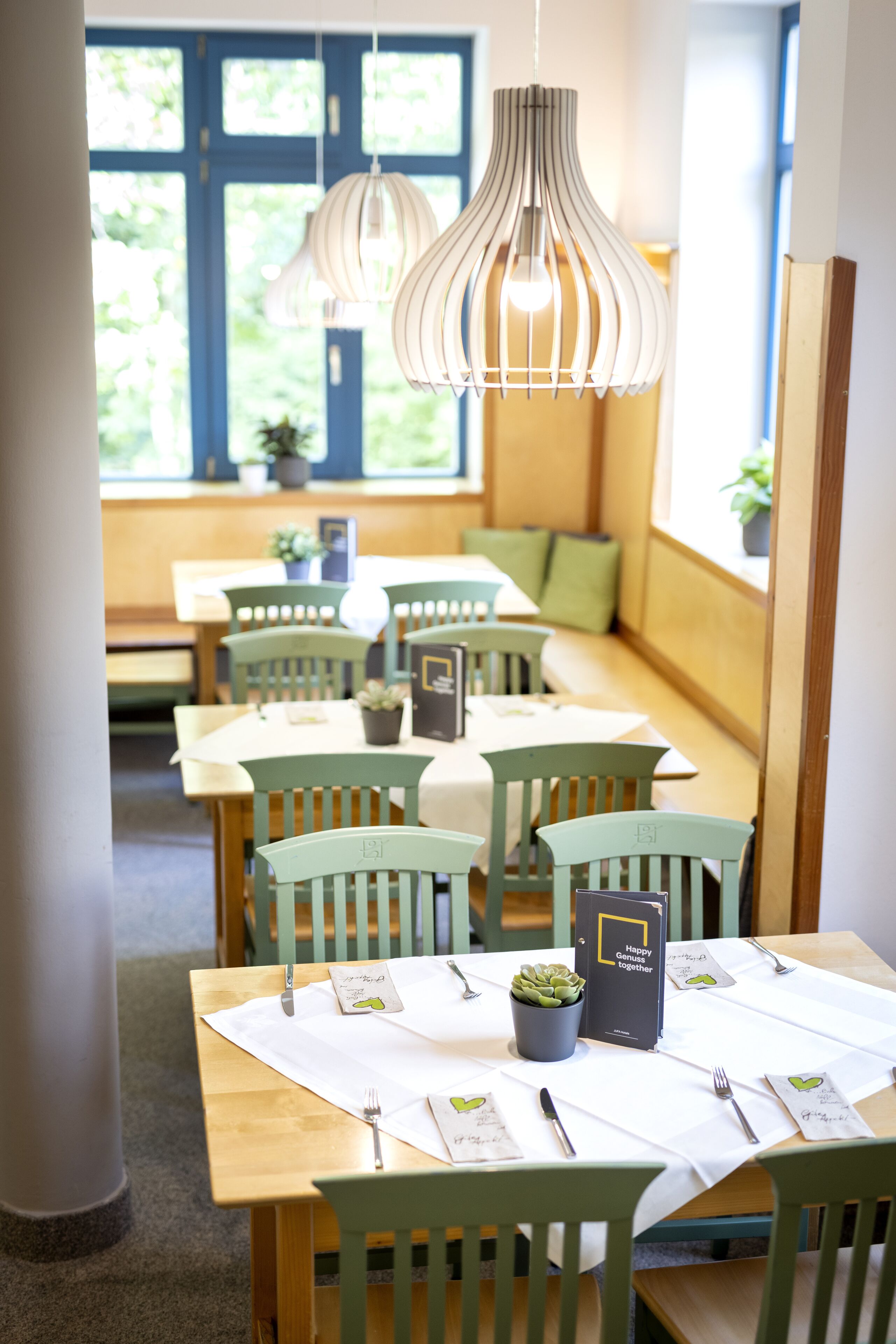 Un espace repas avec des tables et des chaises en bois, des luminaires suspendus et une vue sur la verdure à l’extérieur.
