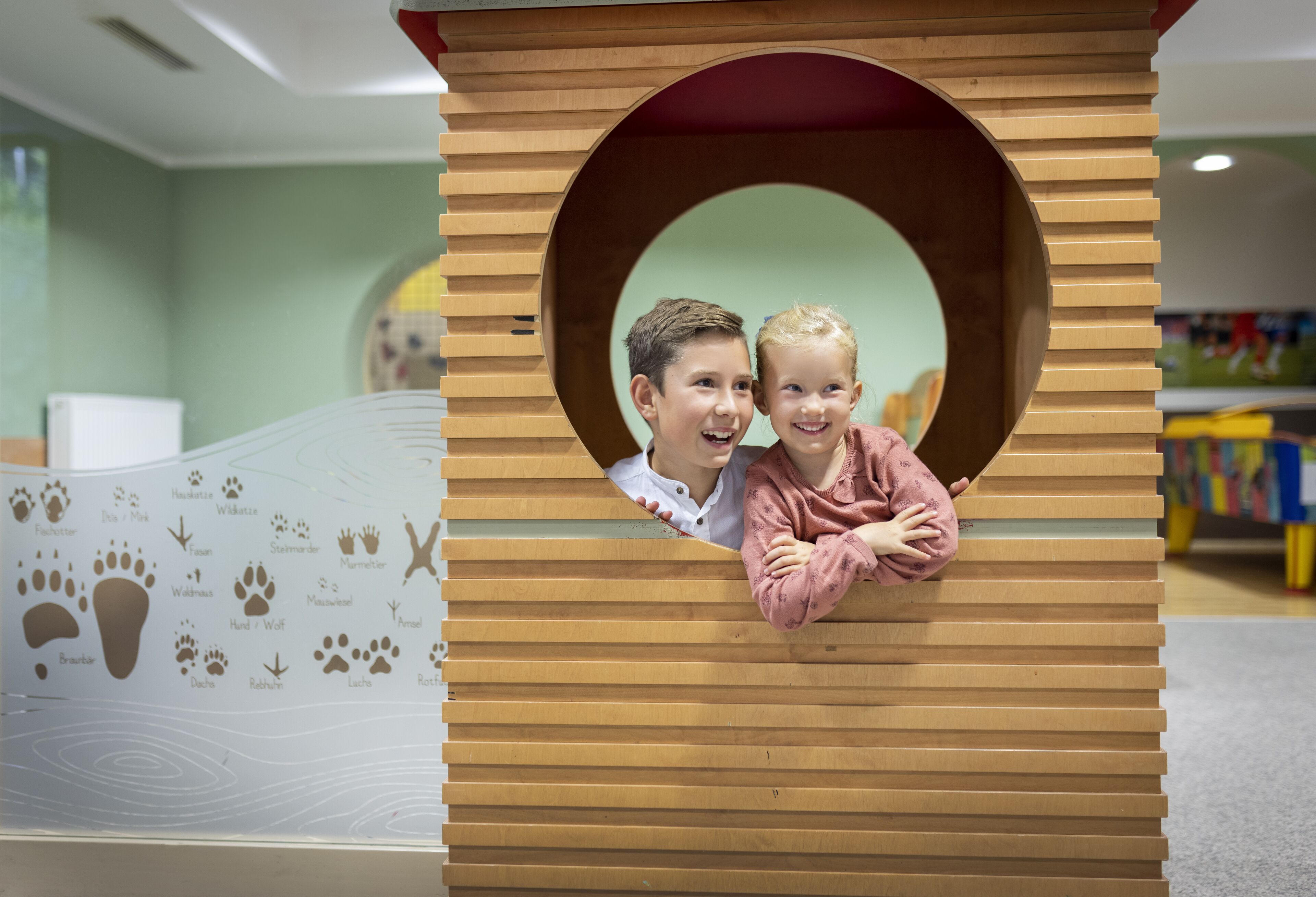 Una casita de juegos de madera con una ventana circular, rodeada por un muro con huellas y nombres de animales.