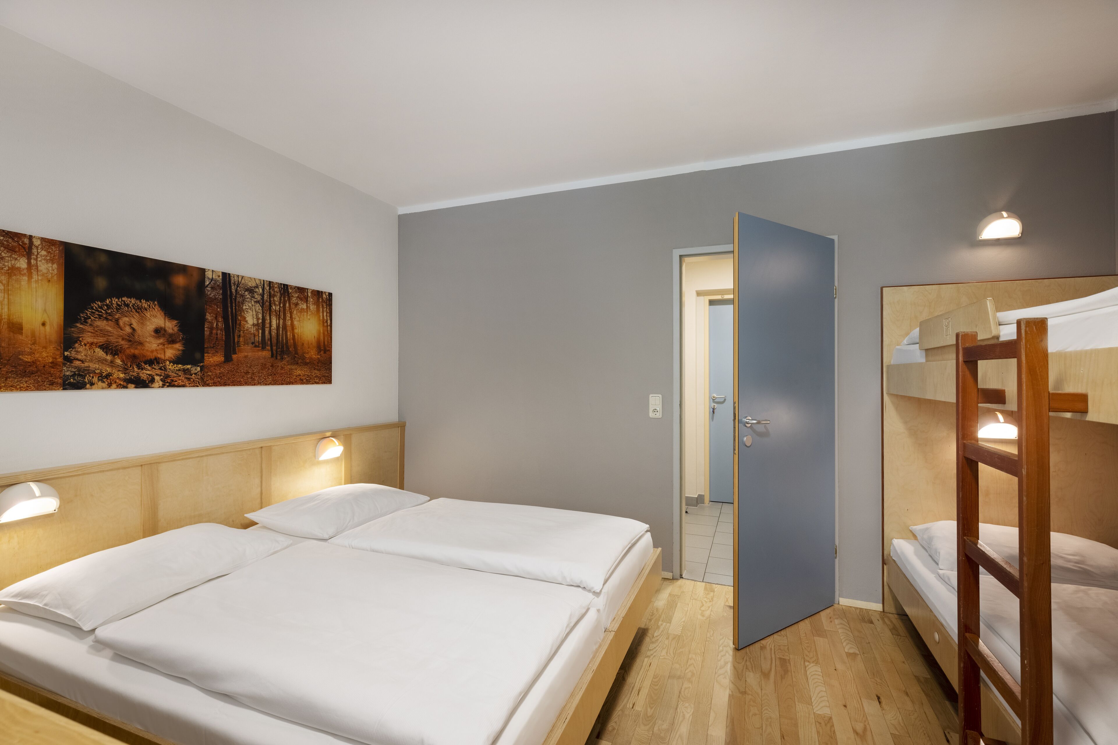 Hotellihuoneessa on suuri sänky, pienempi sänky ja kerrossänky. Seinällä on taulu ja ovi, joka johtaa toiseen huoneeseen.