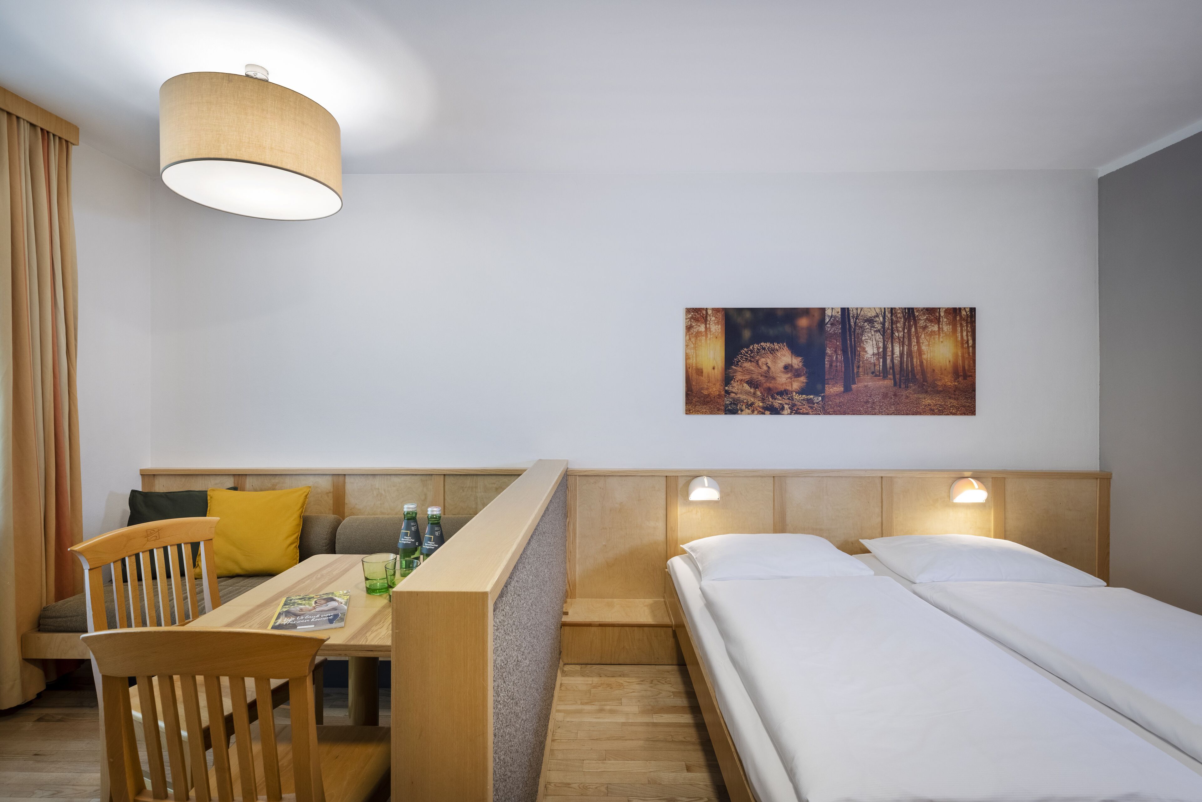 A hotel room with a wooden desk, two beds, a painting on the wall, and a ceiling lamp.