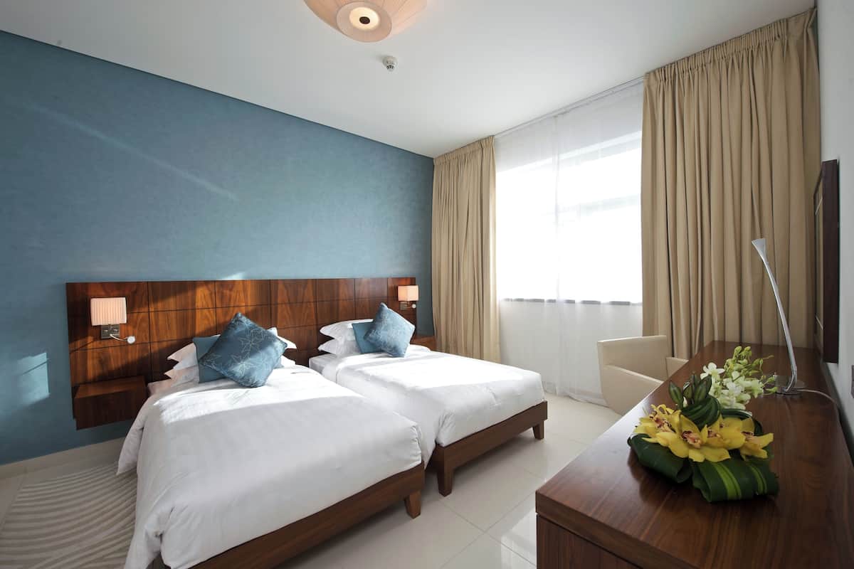 Habitación de hotel con dos camas, cabecera de madera, escritorio con lámpara y ventana con cortinas.