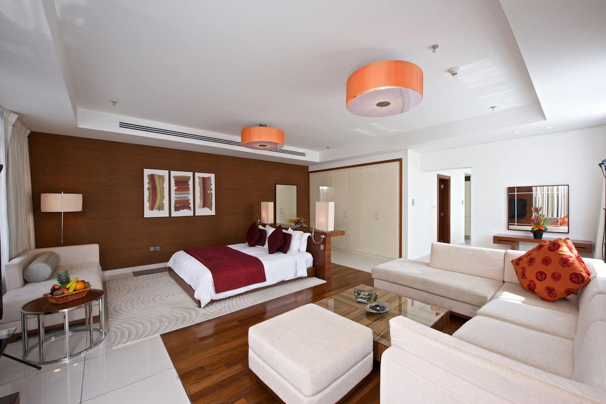 Una habitación de hotel moderna con una cama grande, un sofá, una mesita de centro y una mesa de comedor. Se observan dos lámparas colgantes, un televisor montado en la pared y un ventanal amplio.