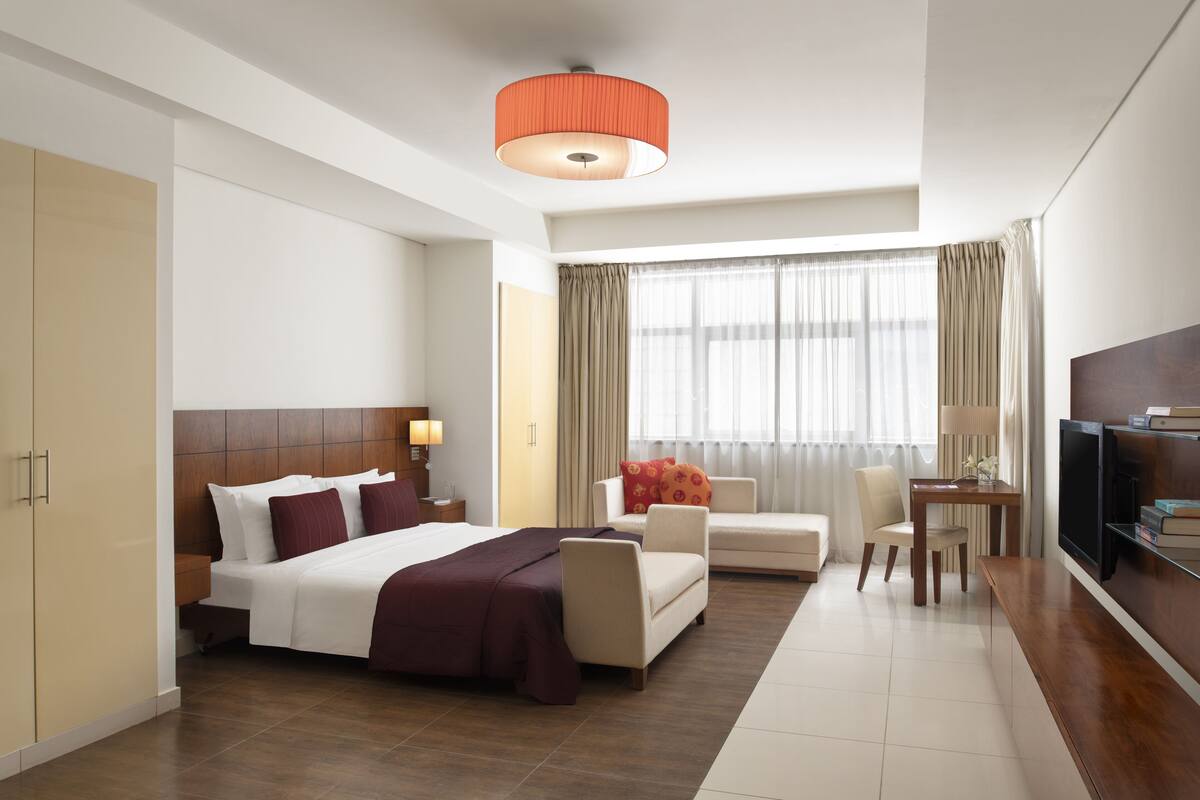Habitación de hotel con cama, escritorio, silla, televisión y un amplio ventanal con cortinas.