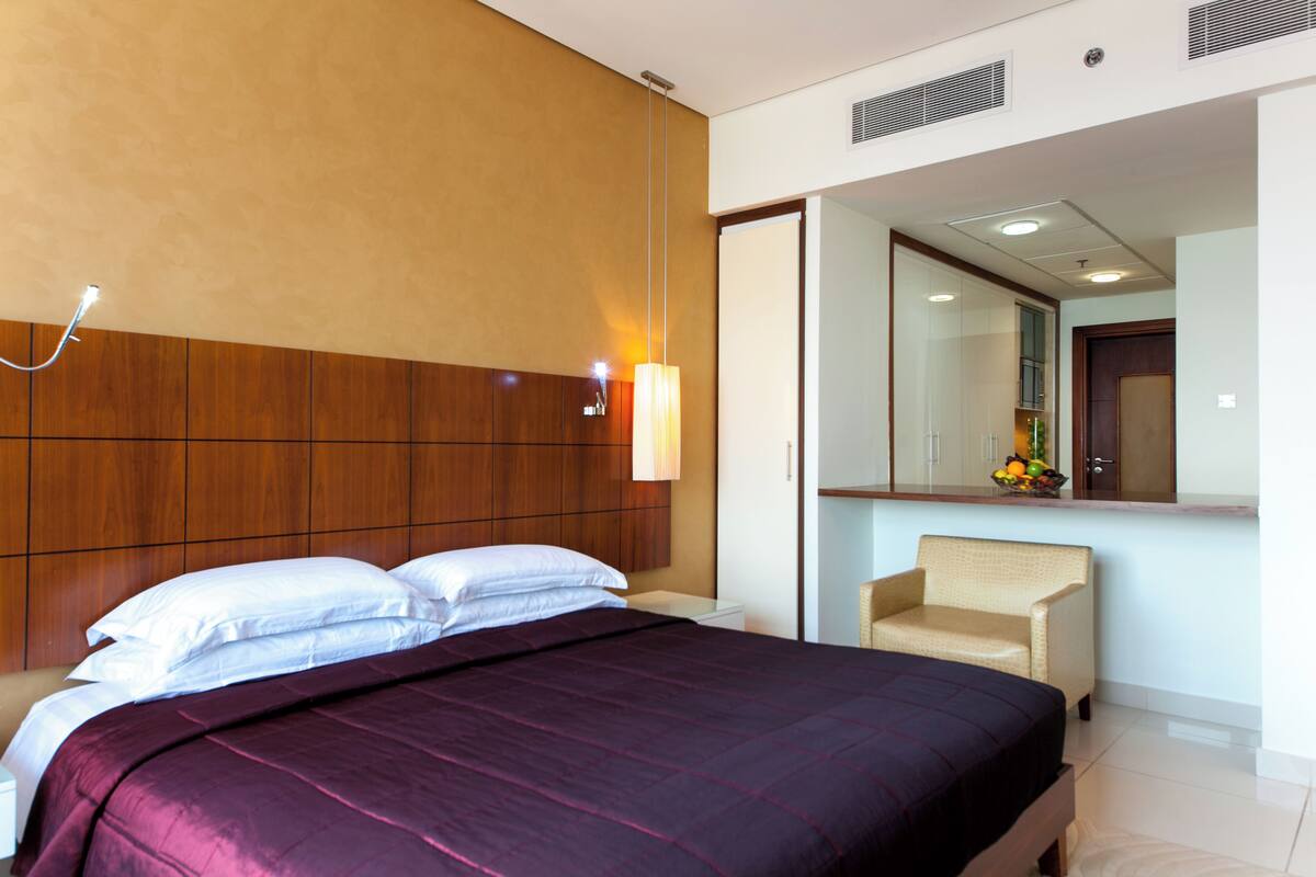 Una habitación de hotel con una cama grande, una mesita de noche, una silla, un espejo y una lámpara fijada a la pared.