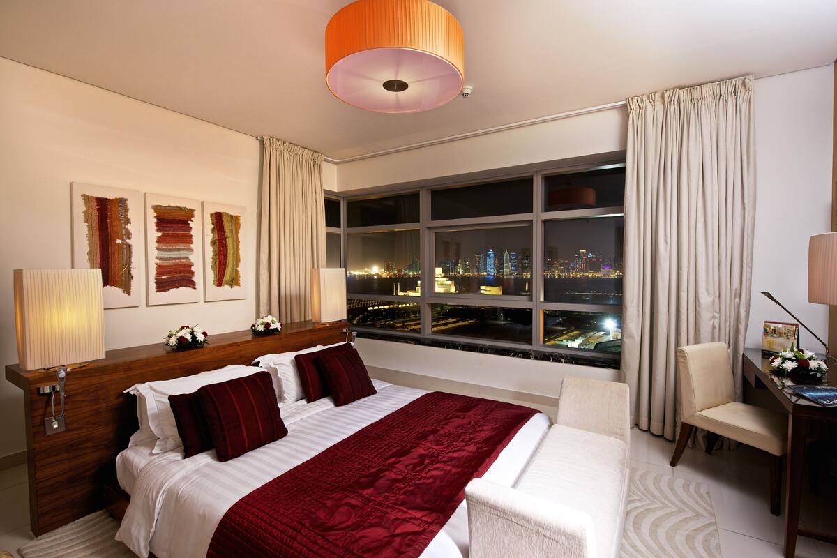 Habitación de hotel con una cama grande, un escritorio, una silla y vistas a un paisaje urbano nocturno.