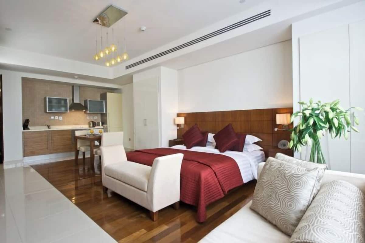 Una habitación de hotel moderna con piso de madera, una cama grande con ropa de cama roja, un sofá blanco, un comedor con mesa y sillas, y una planta grande.