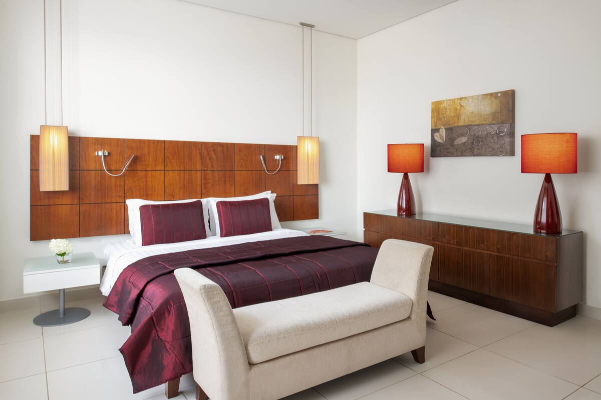 Habitación de hotel moderna con cabecera de madera, una cama grande con ropa de cama roja y blanca, dos lámparas de noche, un aparador de madera y una mesita redonda con un florero.