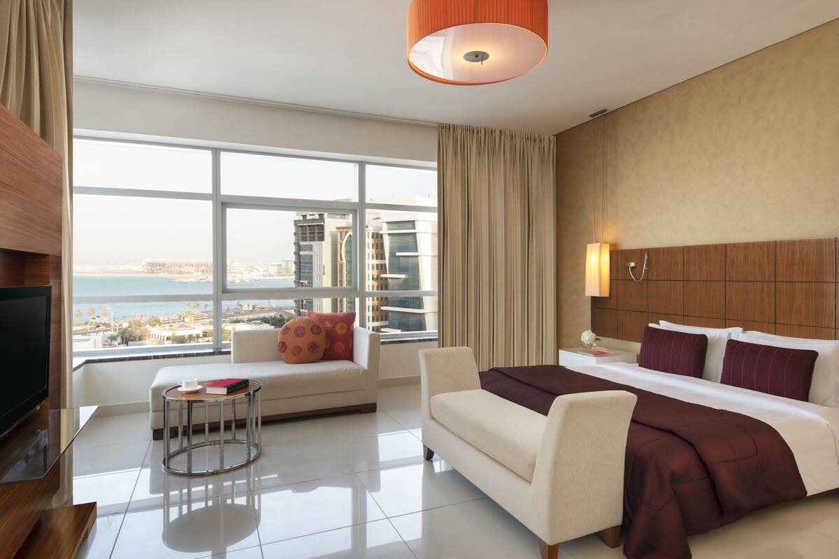 Una habitación de hotel moderna con un amplio ventanal que ofrece vistas a la ciudad, una cama con cabecera, un sofá y un televisor.