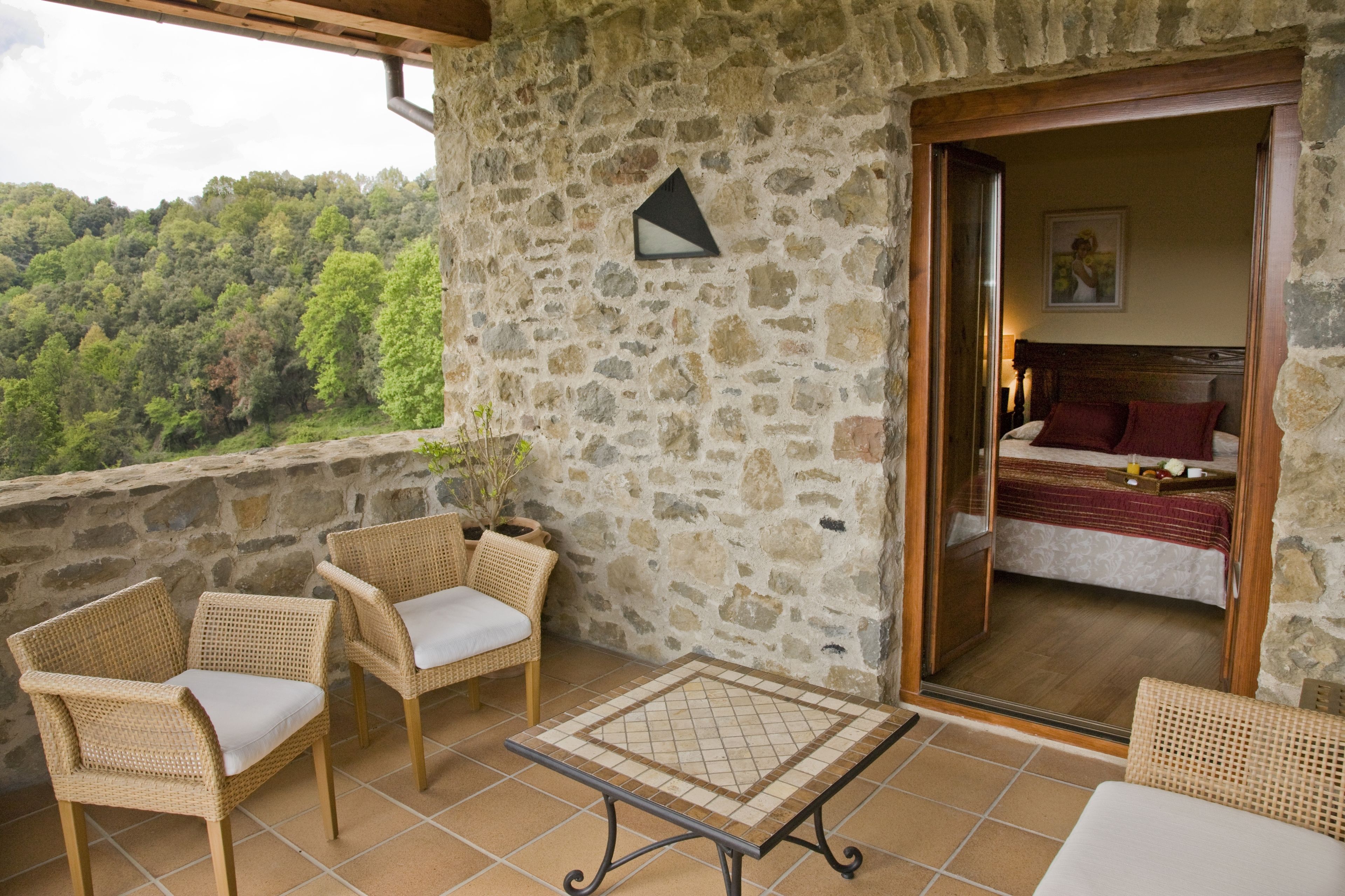 A balcony with wicker furniture, a stone wall, and a bed with a red pillow visible through an open door.
