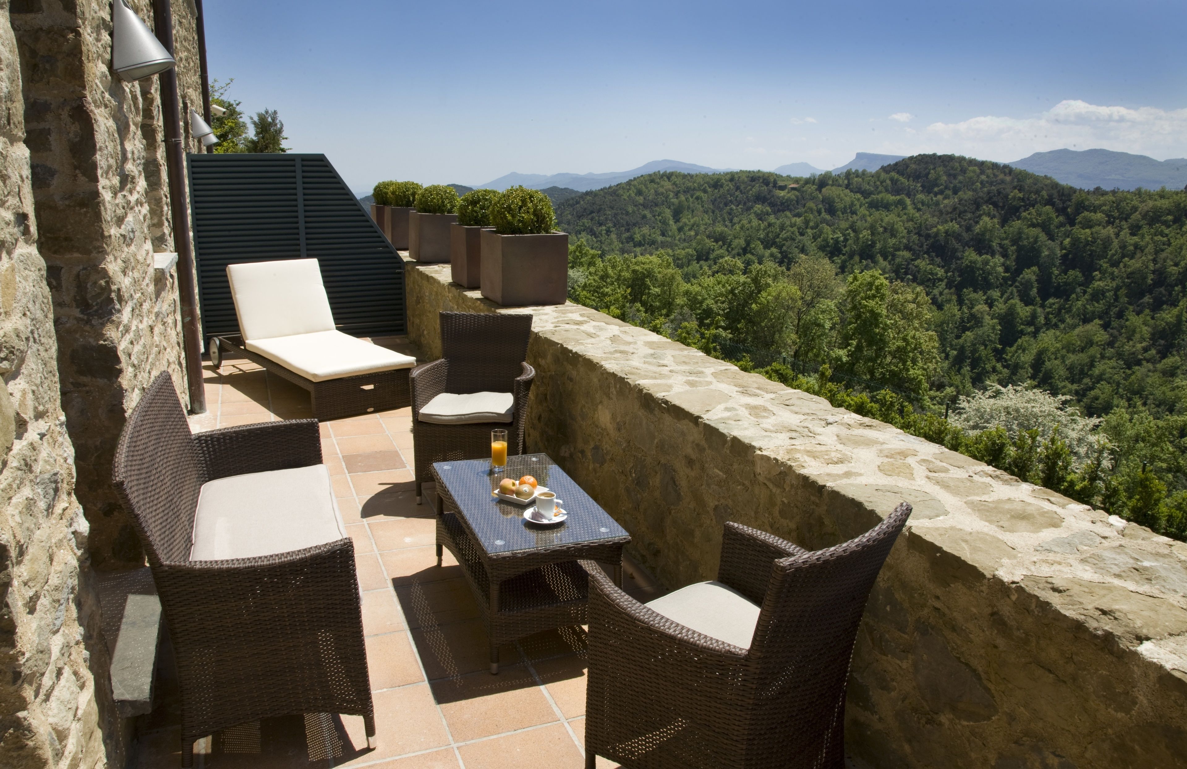 Una terraza con mobiliario de mimbre, una mesa con bebidas y pasteles, y vistas a una cadena montañosa boscosa.