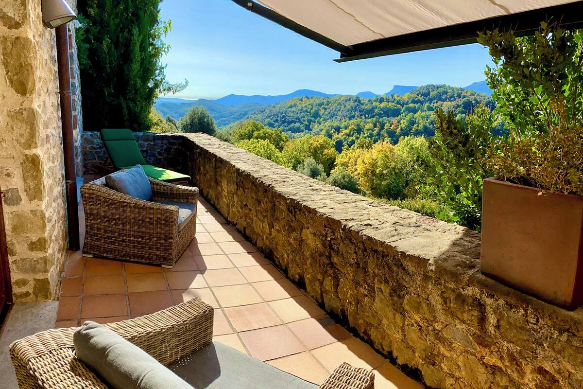 A terrace with wicker furniture, a stone wall, and a view of a forested valley.