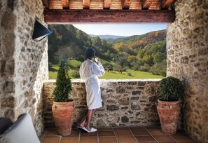 Un balcon aux murs de pierre, agrémenté de pots en terre cuite, doté d’un plafond en bois et offrant une vue sur une vallée verdoyante.