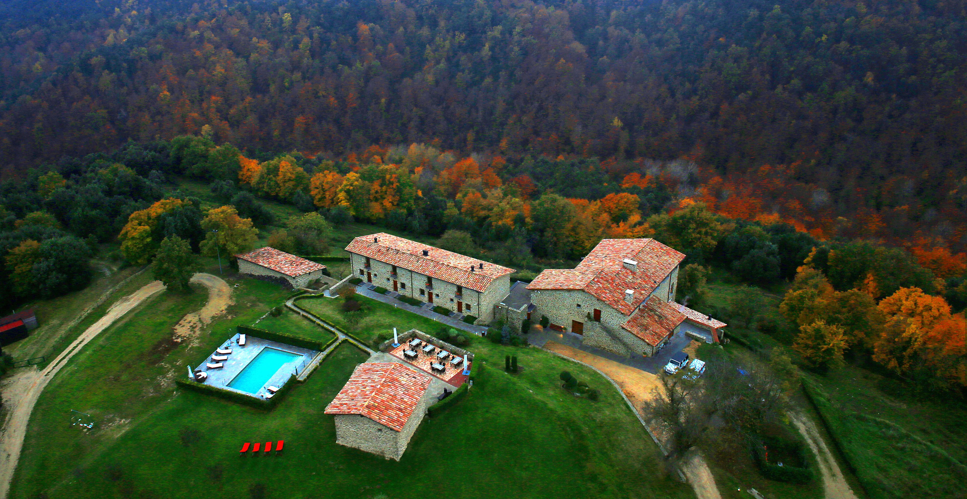An aerial view of a stone building with a swimming pool, surrounded by autumn foliage.