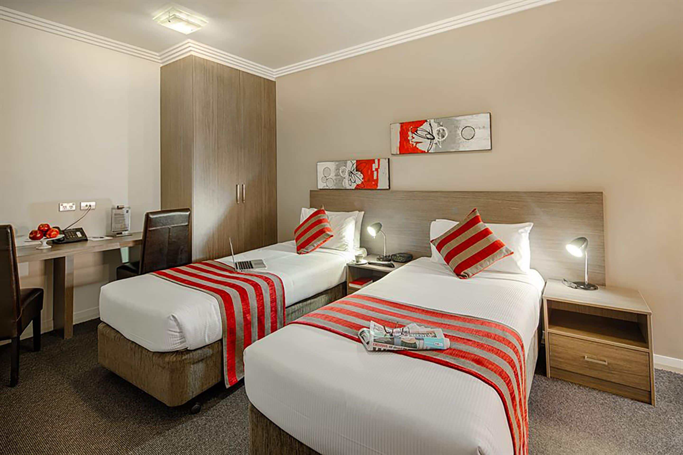 A hotel room with two beds, a desk, a wardrobe, and artwork on the walls.