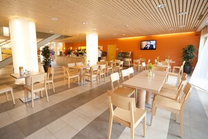 Um espaço amplo e interno, com várias mesas e cadeiras de madeira, uma televisão fixada na parede e uma escada ao fundo.