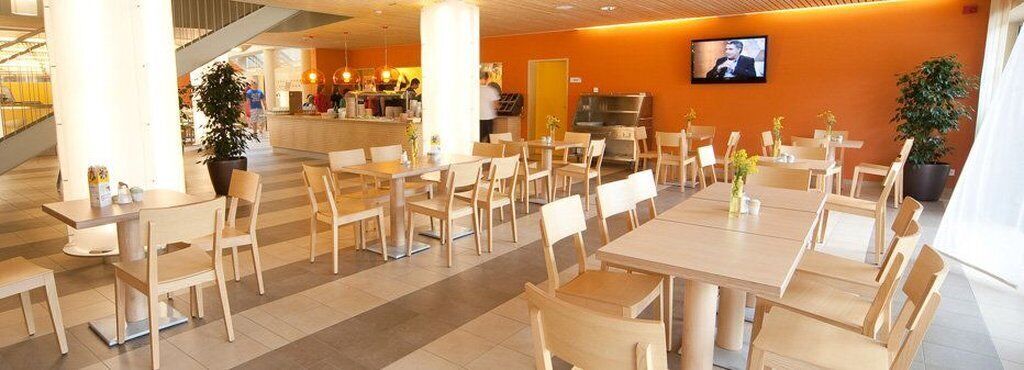 Un moderno vestíbulo de hotel con varias mesas y sillas de comedor, una zona de recepción y una pantalla de televisión en la pared.