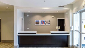 A reception area with a modern design, featuring a reception desk, a wall-mounted security camera, and framed pictures. - Motel 6 Headingley, MB - Winnipeg West (Headingley)