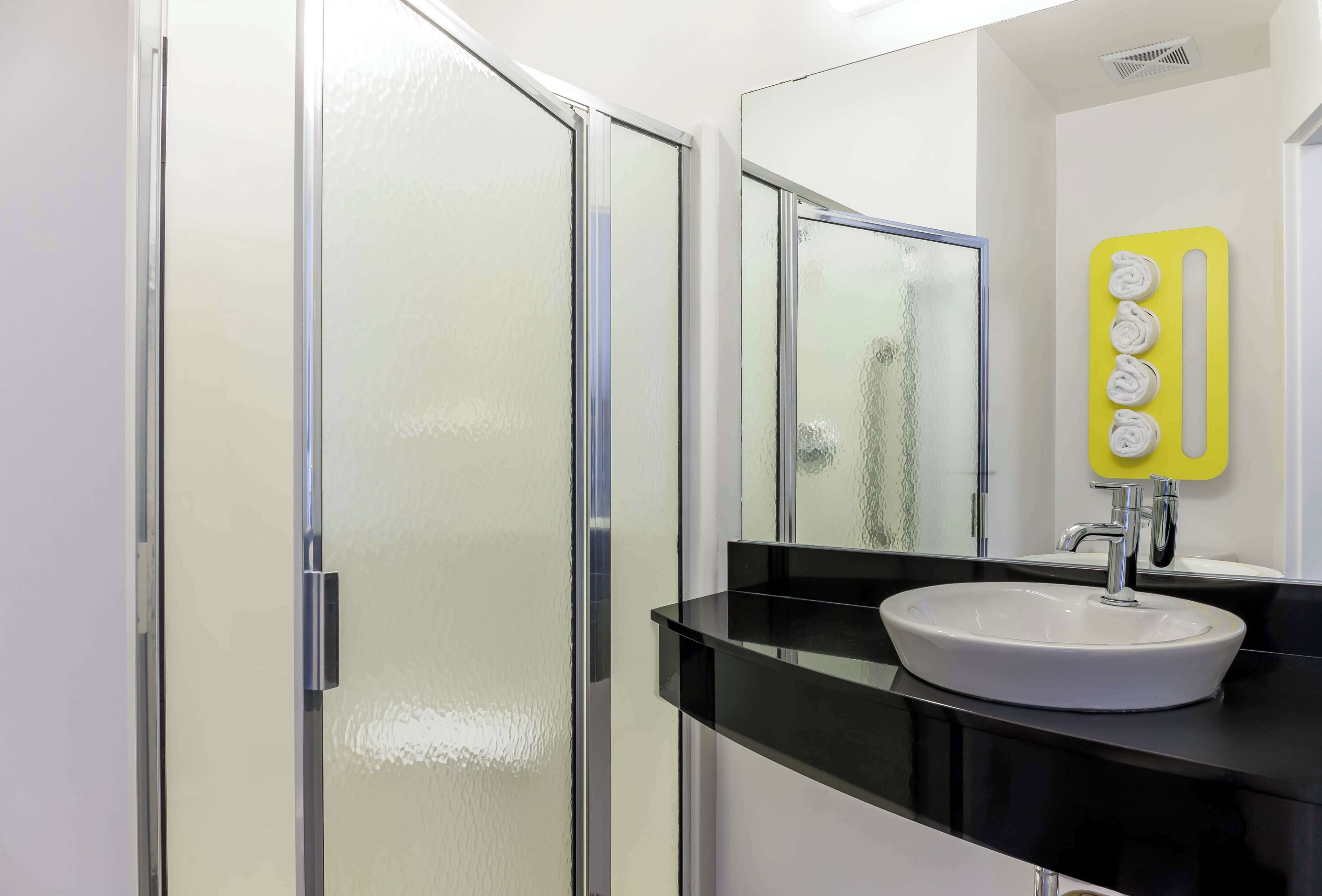 Un baño moderno con cabina de ducha de vidrio, un lavamanos blanco con encimera negra y un espejo con soporte para toallas amarillo.