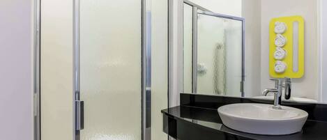 Un baño moderno con cabina de ducha de vidrio, un lavamanos blanco con encimera negra y un espejo con soporte para toallas amarillo.
