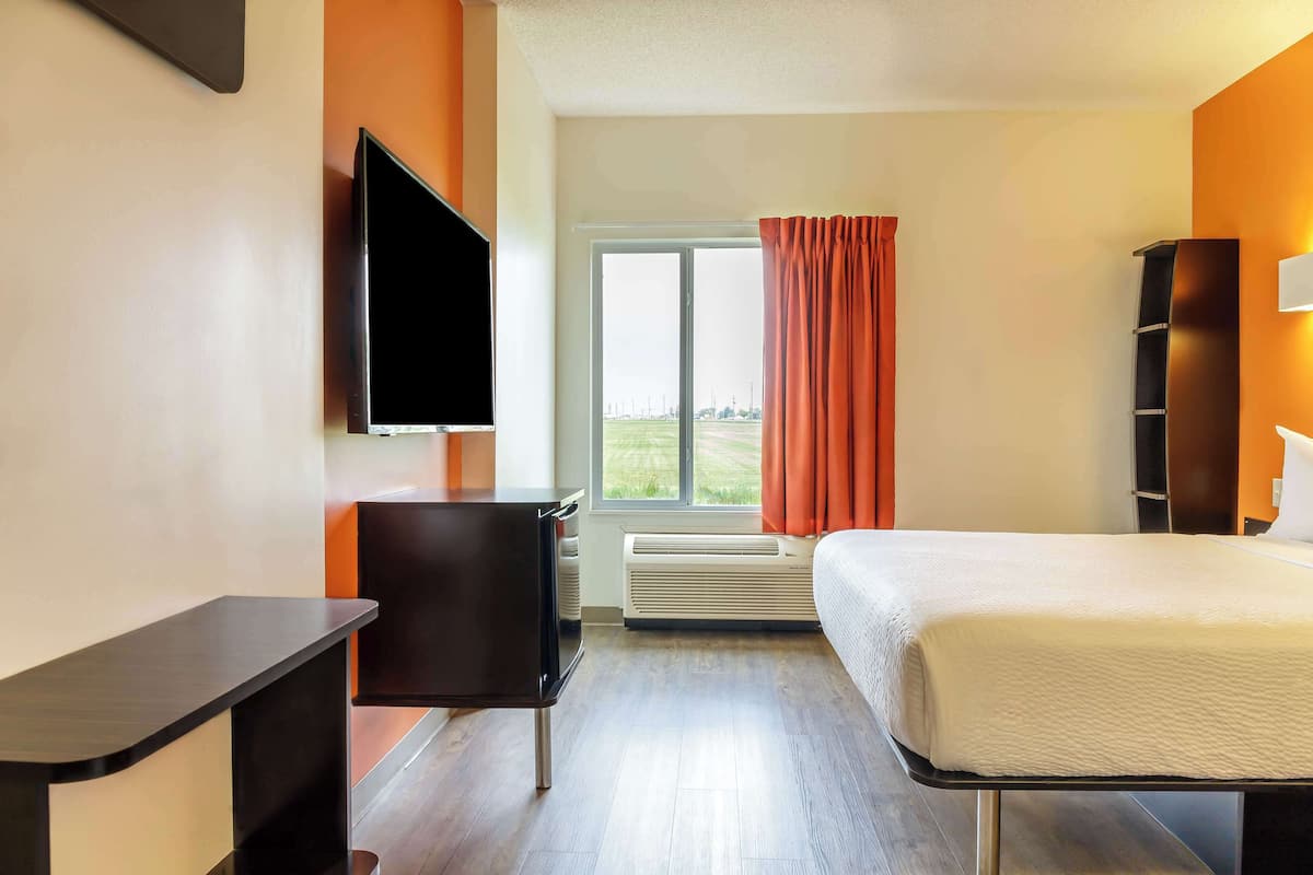 A hotel room with a bed, a desk, a chair, a TV, a window with curtains, and a heater.