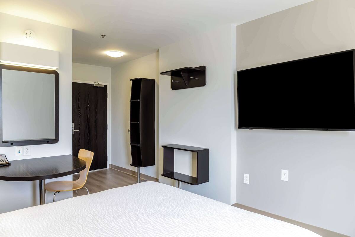 A hotel room with a bed, a desk with a chair, a TV mounted on the wall, and a wardrobe.