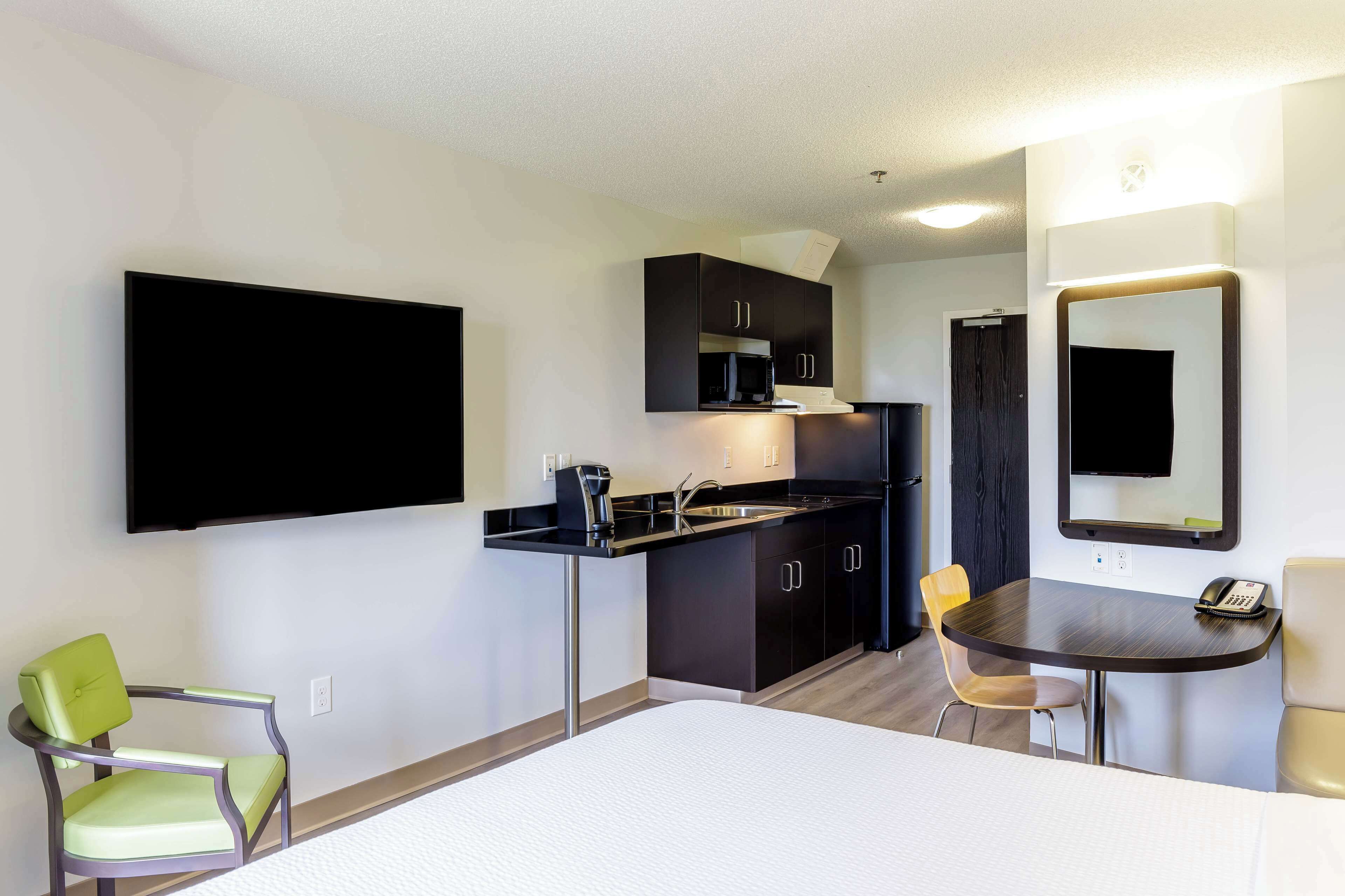 A compact hotel room with a kitchenette, a bed, a single chair, a small table, and a mirror.