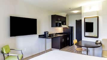 A compact hotel room with a kitchenette, a bed, a single chair, a small table, and a mirror.