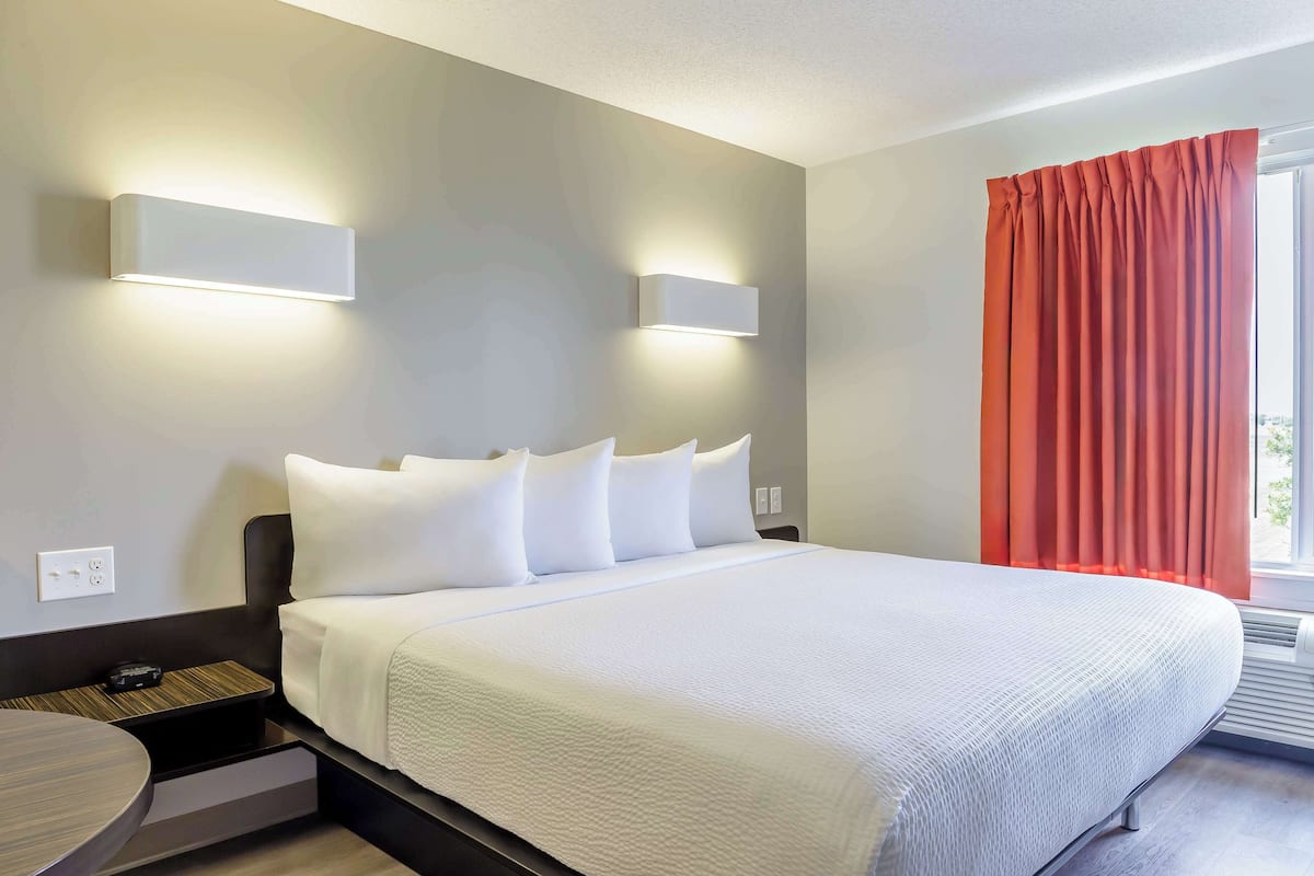A modern hotel room with a large bed, white bedding, two wall-mounted lights, a bedside table with a lamp, and a window with red curtains.