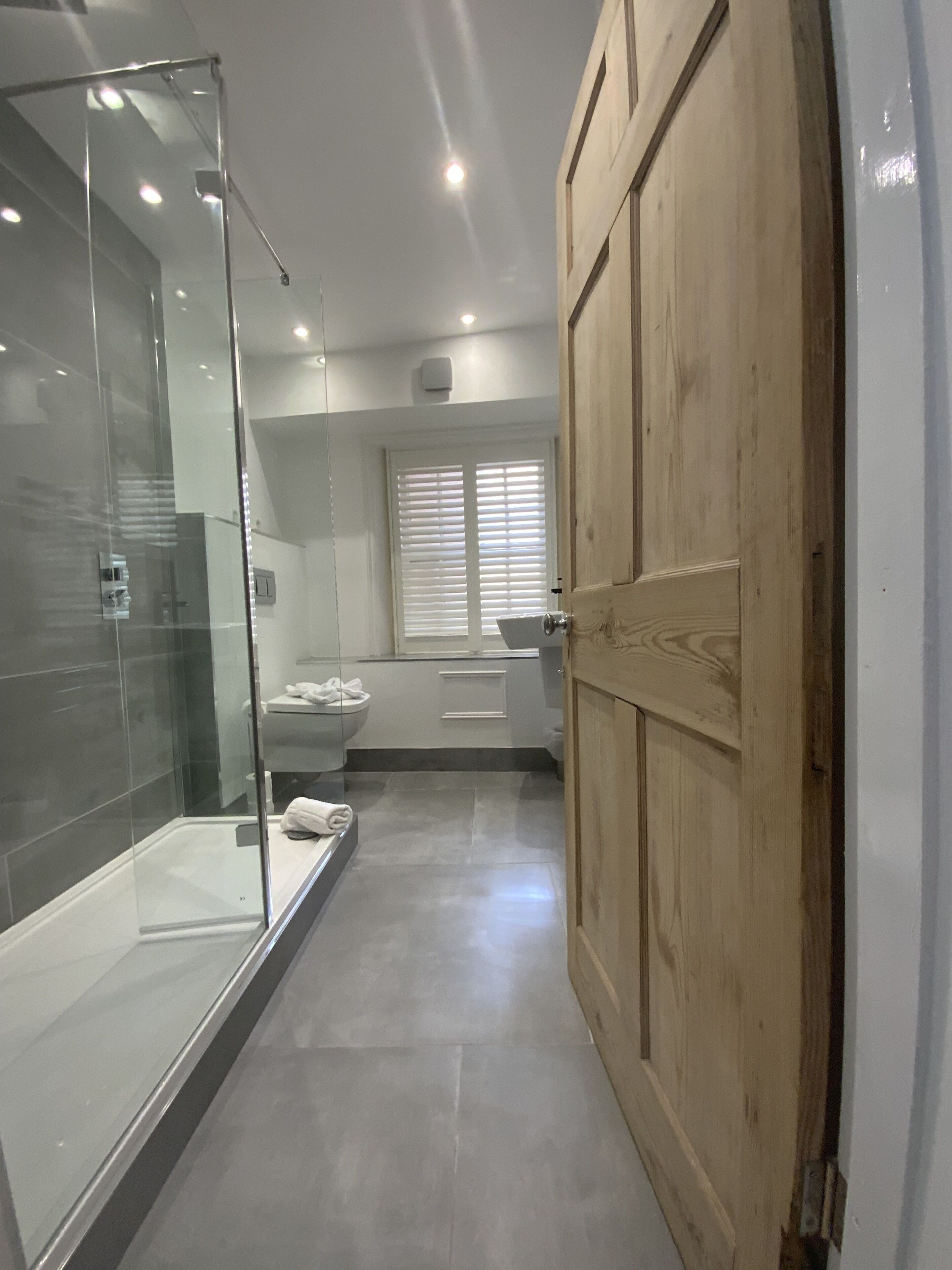 Un baño moderno con cabina de ducha de vidrio, puerta de madera y ventana con persianas.