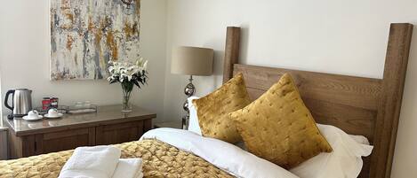 A neatly made bed with a gold quilted cover, white pillows, and a wooden headboard. A nightstand with a lamp and a vase of flowers is beside the bed.