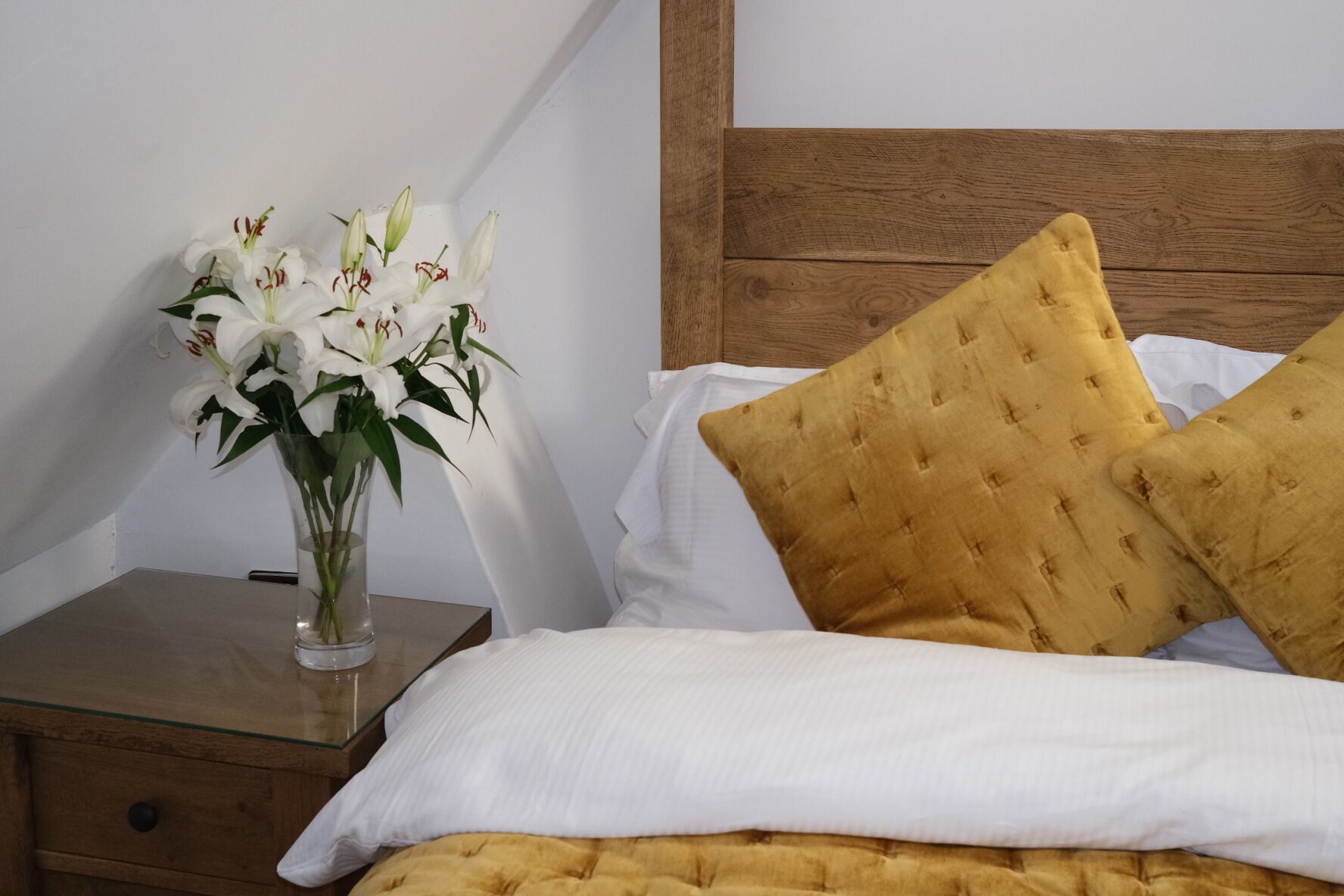 Una cama bien hecha con almohadas blancas y de color mostaza, un florero de vidrio transparente con flores blancas sobre una mesita de noche de madera, y un cabecero de madera.