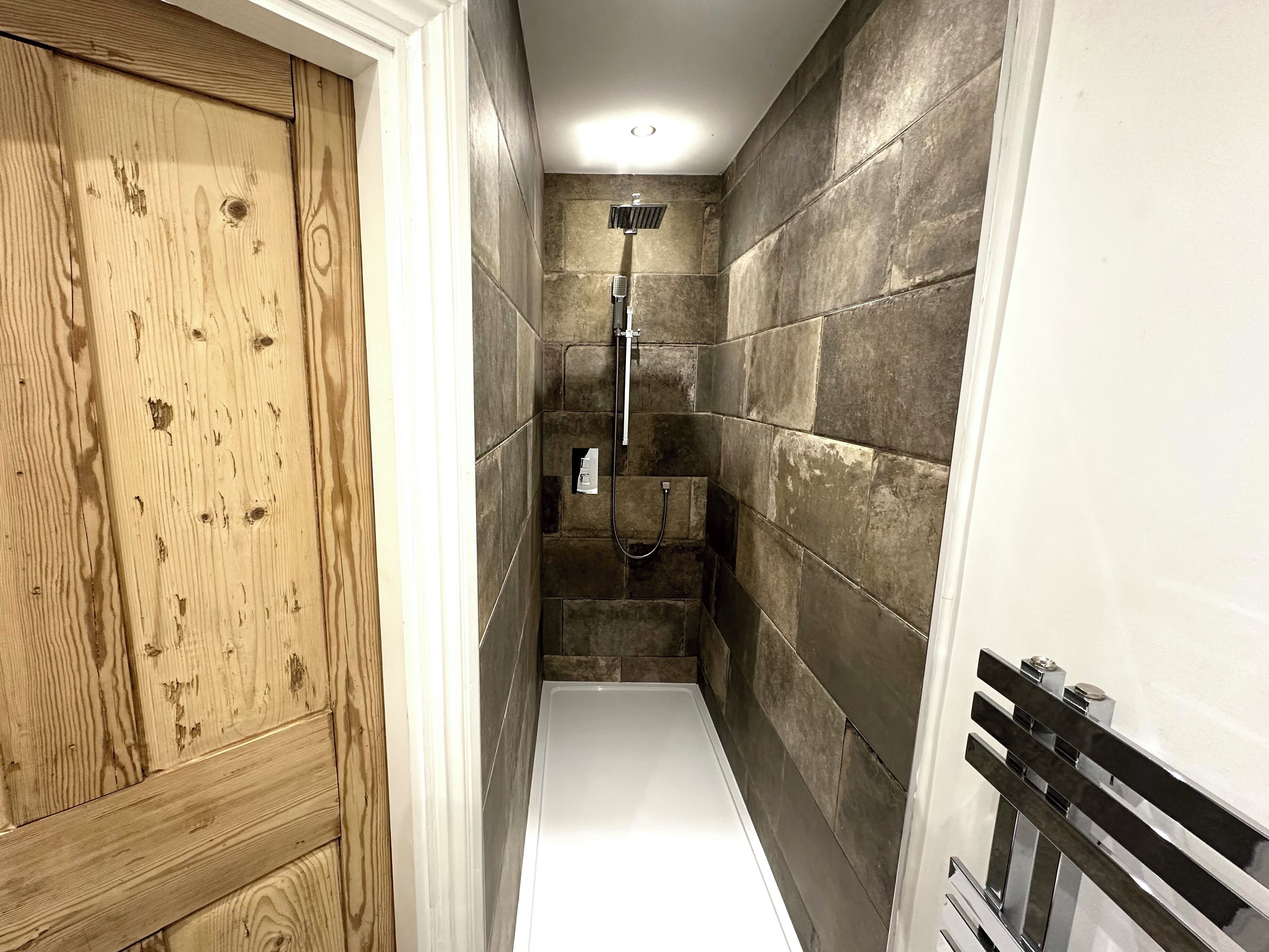Une petite salle de bain avec une porte en bois, des murs carrelés, une douche et un sol blanc.
